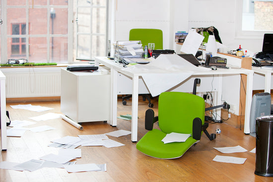 Office with white walls, desk, and cabinets, and a light wood floor. A lime green office chair is on both sides of the desk, one is turned on its back., Papers are strewn around the floor and desk. There are also a few bottles on desk.