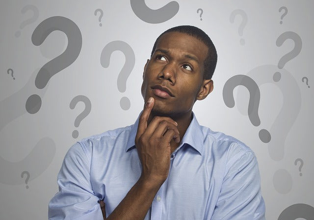 African-American man looking up and to his right with hand on his face, looks like he's thinking. He's wearing a light blue button-up shirt. The wall behind him is white with random grey question marks on it.