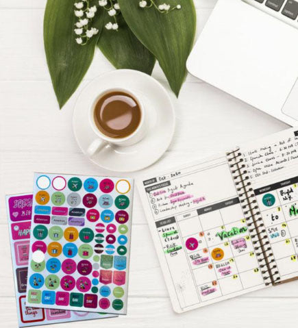 Two sheets of colorful round and rectangular stickers, a cup of coffee on a saucer, a laptop, a lily of the valley plant, and an open planner with writing and stickers inside are all sitting on a white wooden surface.